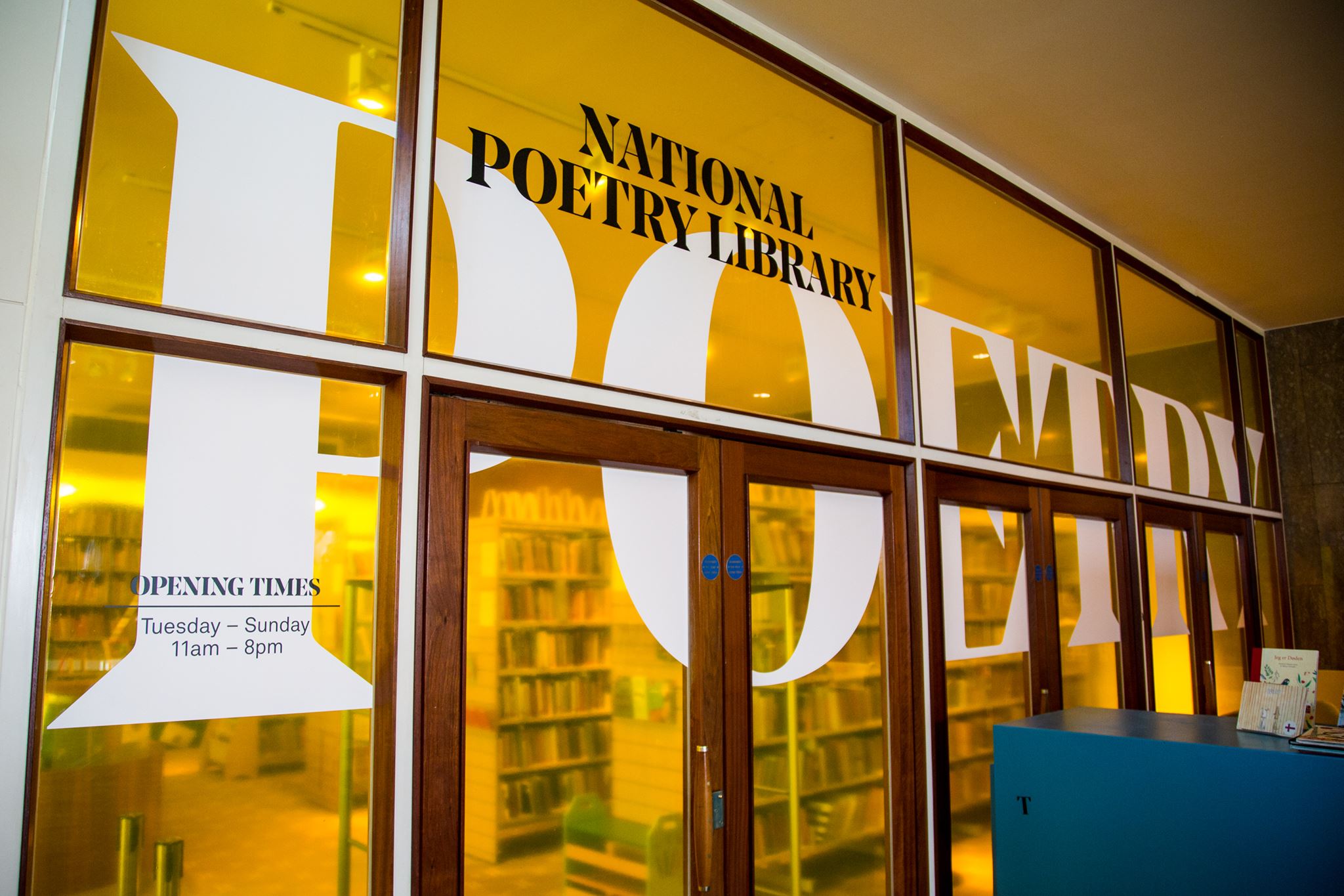 Photograph of the entrance to the National Poetry Library in London. The picture shows the glass doors in a wall of coloured yellow glass with the word poetry written in large white letters across the whole length of the wall.